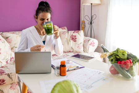 An adult nutritionist doctor in a white coat at the table with vegetables waving her hand at a laptop has a video call with the patient in the clinic interior.の写真素材