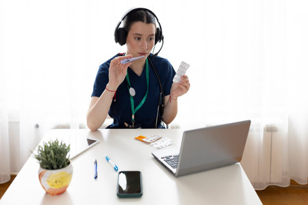 Young doctor woman wears headphones sit at desk look at laptop, provide professional medicare to clinic patient remotely using videocall telemedicine platform application. Medicineの写真素材