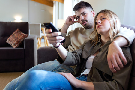 Portrait of beautiful young bored couple watching TV in the sofa at home.