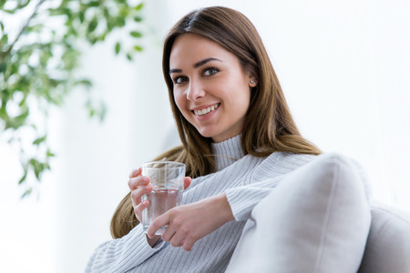 Portrait of beautiful young woman looking at camera while holding glass of water at home.