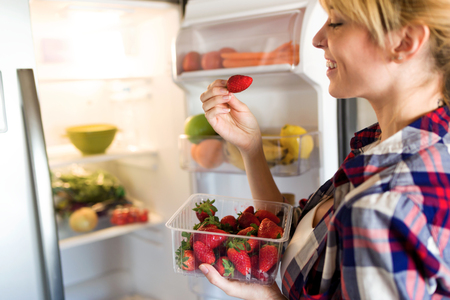Shot of pretty young woman picking strawberries of fridge in the kitchen.の写真素材