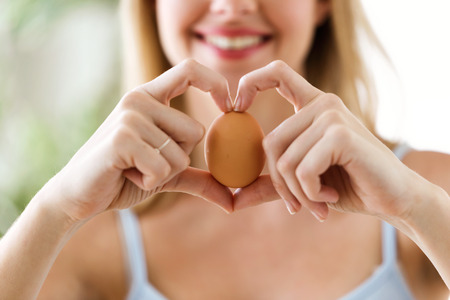 Shot of beautiful young woman showing brown chicken egg with hands in a heart shape at home.の写真素材