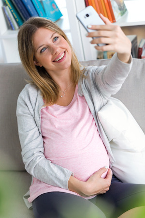 Portrait of beautiful pregnant woman taking a self-portrait with her smartphone at homeの写真素材