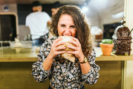 Portrait of beautiful young woman eating hamburguer in the food truck.の写真素材