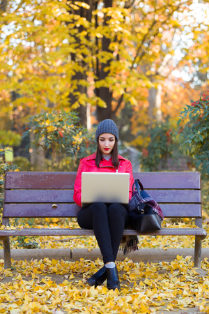 Portrait of beautiful young woman sitting in a bench and using her laptop in autumn.の写真素材