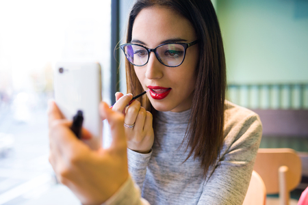 Portrait of beautiful young woman doing make-up with her lipstick and mobile phone.の写真素材