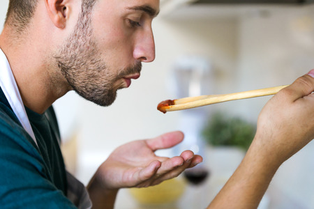 Close up of handsome young man tasting the fried with wooden spoon in the kitchen at home.の写真素材