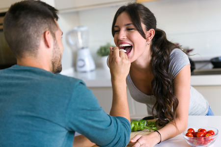 Shot of happy young couple enjoying lunch while handsome man is feeding with yogurt his wife in the kitchen at home.の写真素材