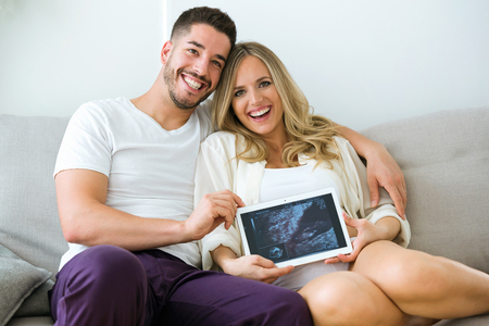 Portrait of attractive happy young man and pregnant smiling woman looking at camera while holding digital tablet and showing baby ultrasound at home.の写真素材