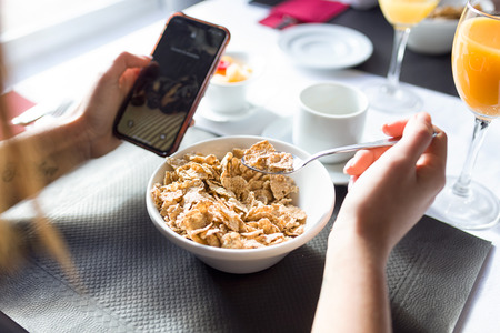 Close-up of young woman having breakfast and texting with her smartphone in dining room of the...