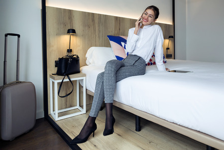 Shot of stylish young businesswoman talking with her mobil phone sitting on the bed at hotel room.の写真素材