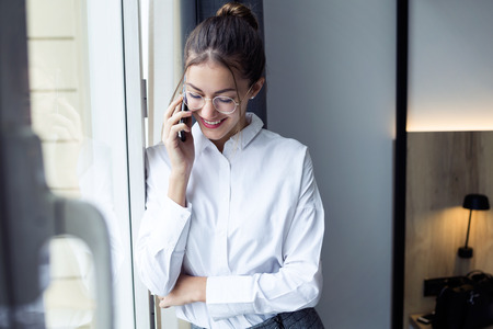 Shot of pretty young businesswoman talking with her smartphone standing near the window at hotel...