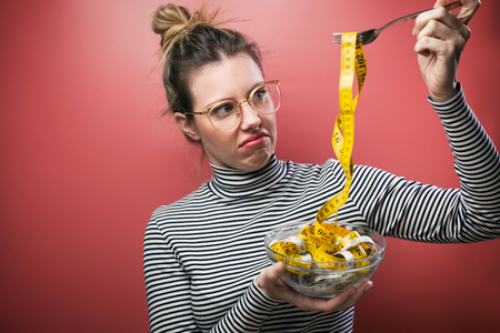 Shot of disgusting woman with eyeglasses holding bowl with measuring tapes while over pink background.の写真素材