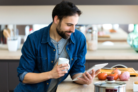 Shot of handsome young man using his digital tablet while drinking coffee in the kitchen at home.の写真素材