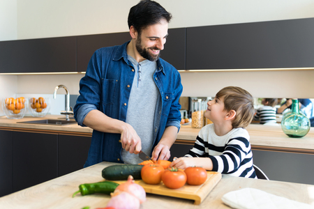 Shot of attractive young father and his little cute son cutting vegetables on wooden table  in the kitchen at home.の写真素材