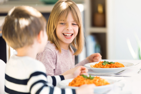 Shot of smiling children having fun while eating spaghetti with tomato sauce in the kitchen at home.の写真素材