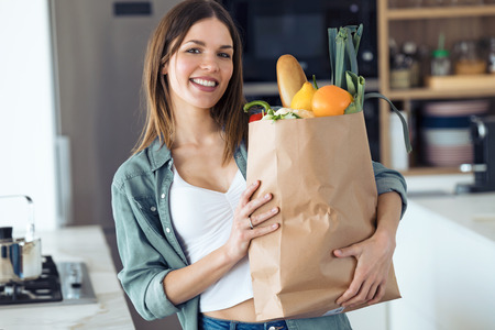 Portrait of beautiful young woman looking at camera while holding shopping bag with fresh vegetables in the kitchen at home.の写真素材