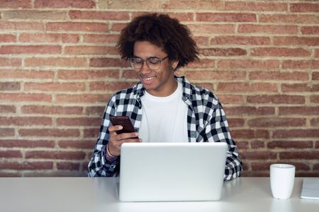Shot of handsome young afro-american man with eyeglasses using his smartphone while working with...