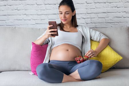 Shot of beautiful pregnant young woman using her mobile phone while eating strawberries on sofa at...