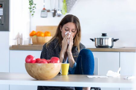 Shot of illness young woman sneezing in a tissue while sitting in the kitchen at home.の写真素材