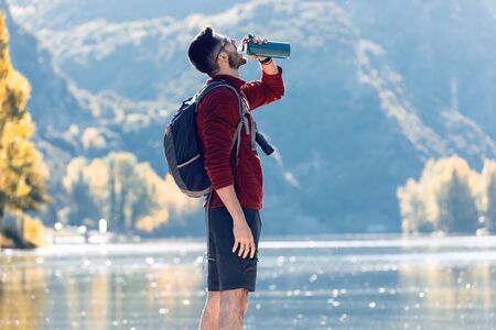 Hiker young man traveler with backpack drinking water while standing in front of lake.の写真素材