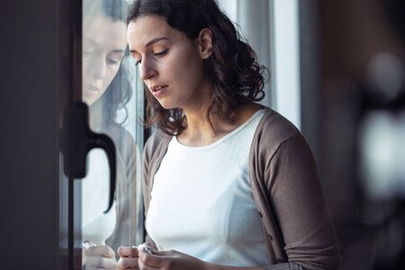 Shot of sad young woman crying while looking through the window at home.