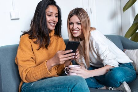 Shot of two pretty young women using mobile phone while drinking coffee on sofa at home.の写真素材