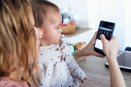 Close-up of young woman using her mobile phone while her baby girl looks at it at home.
