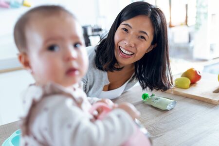 Shot of happy young mother with her baby daughter looking at camera in the kitchen at home.の写真素材