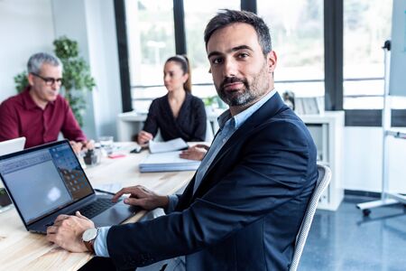 Portrait of handsome businessman is looking at camera while his colleagues are working on coworking...
