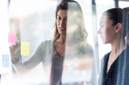 Shot of two business young women working together on wall glass with stickers on co-working space.