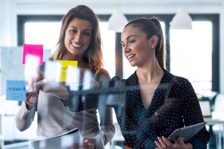 Shot of two business young women working together on wall glass with stickers on co-working space.の写真素材