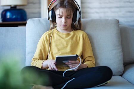 Shot of little girl listening to music with headphones and digital tablet while sitting on sofa at...