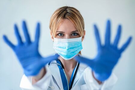 Portrait of female doctor with a medical mask showing hands to camera while wearing a blue nitrile...