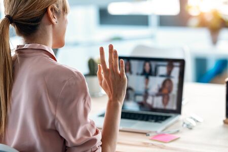 Back view of female employee waving and speaking on video call with diverse colleagues on online briefing with laptop at home.の写真素材