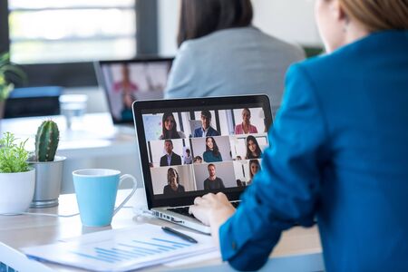 Back view of two business women speaking on video call with diverse colleagues on online briefing with laptop in the office.の写真素材