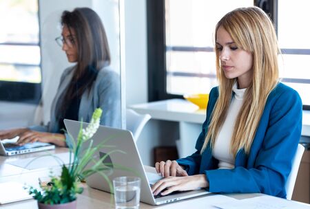 Shot of two business women work with laptops on the partitioned desk in the coworking space. Concept...