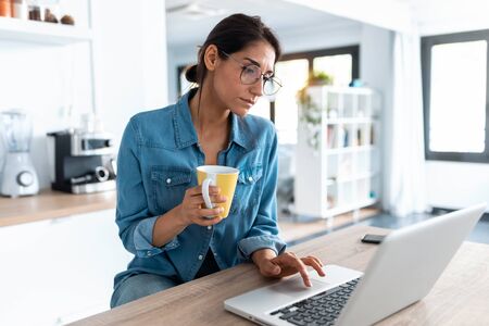 Shot of pretty young woman working with laptop during morning coffee in the kitchen at home.の写真素材