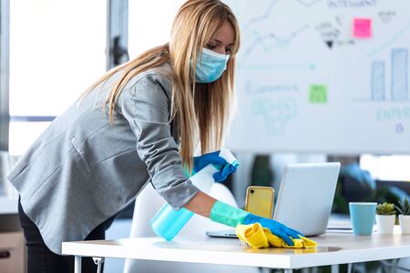Shot of young business woman with face mask and protective gloves cleaning computer desk with spray...