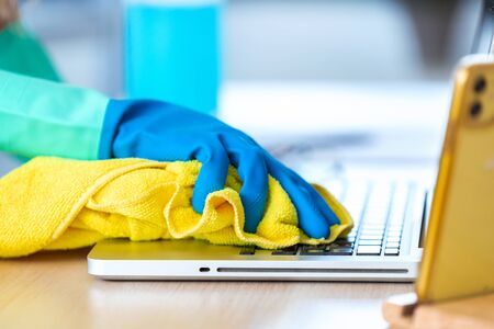 Close-up of woman with protective gloves cleaning the laptop with spray and rag in office.