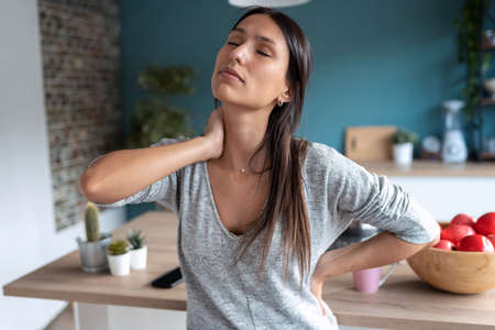 Shot of tired young woman suffering neck pain while sitting on the stool in the kitchen at home.の写真素材
