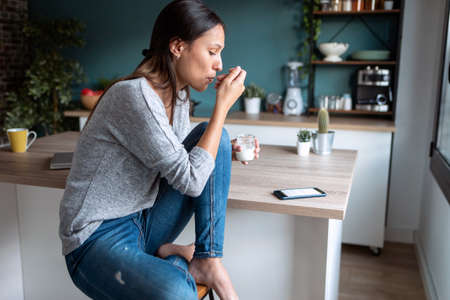 Shot of smiling young woman using her mobile phone while eating yogurt in the kitchen at home.の写真素材