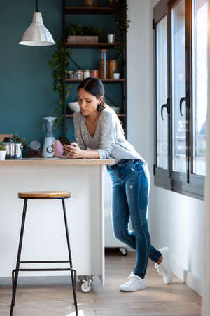 Shot of smiling young woman using her mobile phone while eating yogurt in the kitchen at home.の写真素材