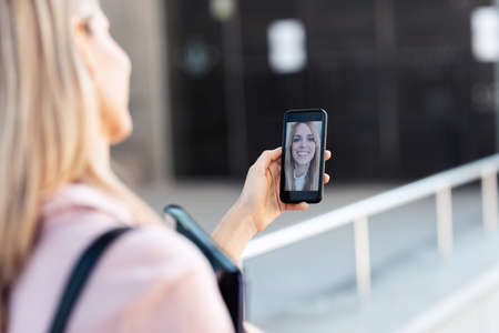 Shot of back view of woman having video conversation with pretty young business woman while staying in the street.の写真素材
