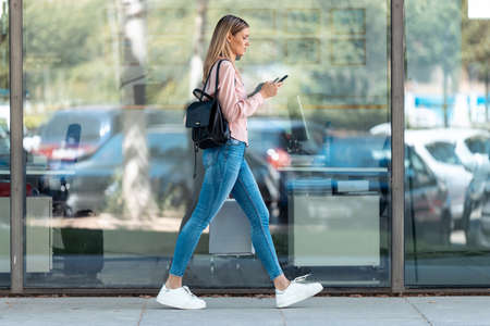 Shot of beautiful blonde female student using her smartphone while walking on campus of university.の写真素材