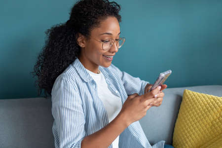 Shot of smiling young african american woman using her mobile phone while sitting on sofa at home.
