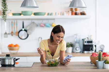 Shot of beautiful young woman using her mobile phone while eating a salad in the kitchen at home.の写真素材
