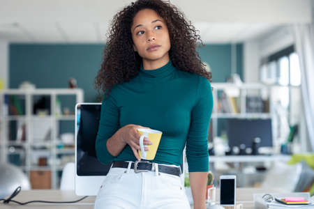Portrait of pensive young woman looking outside while holding a cup of coffee in the office.