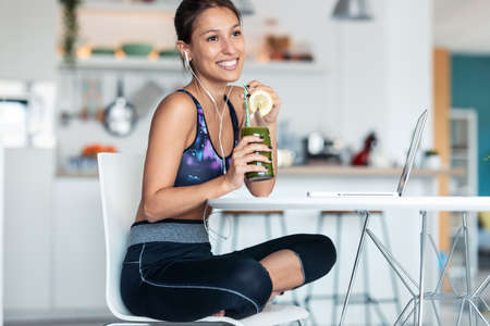 Shot of sporty young woman drinking detox juice while working with her laptop after session of...