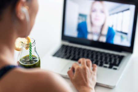 Close-up of sporty beautiful woman having an online video call via laptop computer while drinking detox juice in the kitchen at home.の写真素材